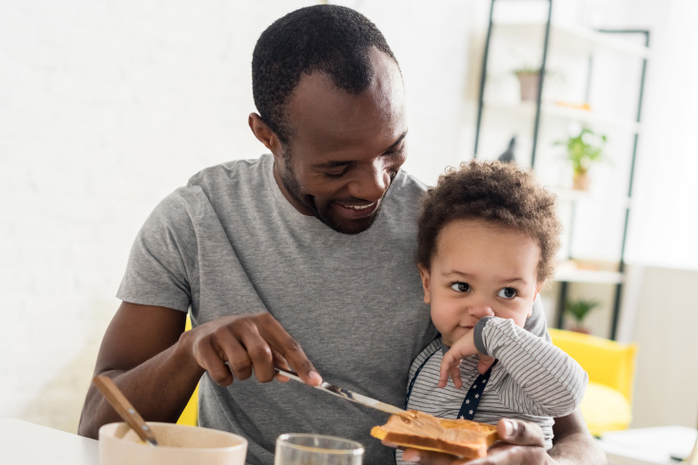 Father with young son on his lap spreading peanut butter on toast.