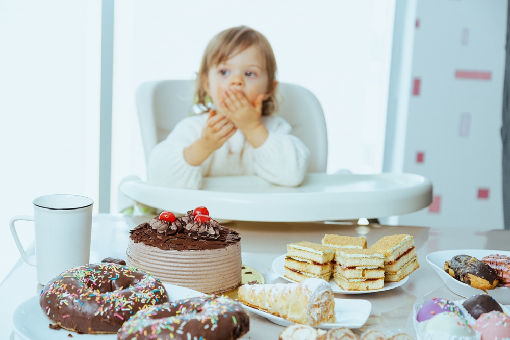 Toddler sitting in a highchair looking at an assortment of cakes and sweets on a table.